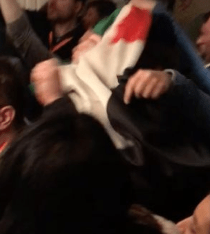 A protester smothers a Jewish man wearing a yamulkah, with a Palestinian flag, attacking him from behind.